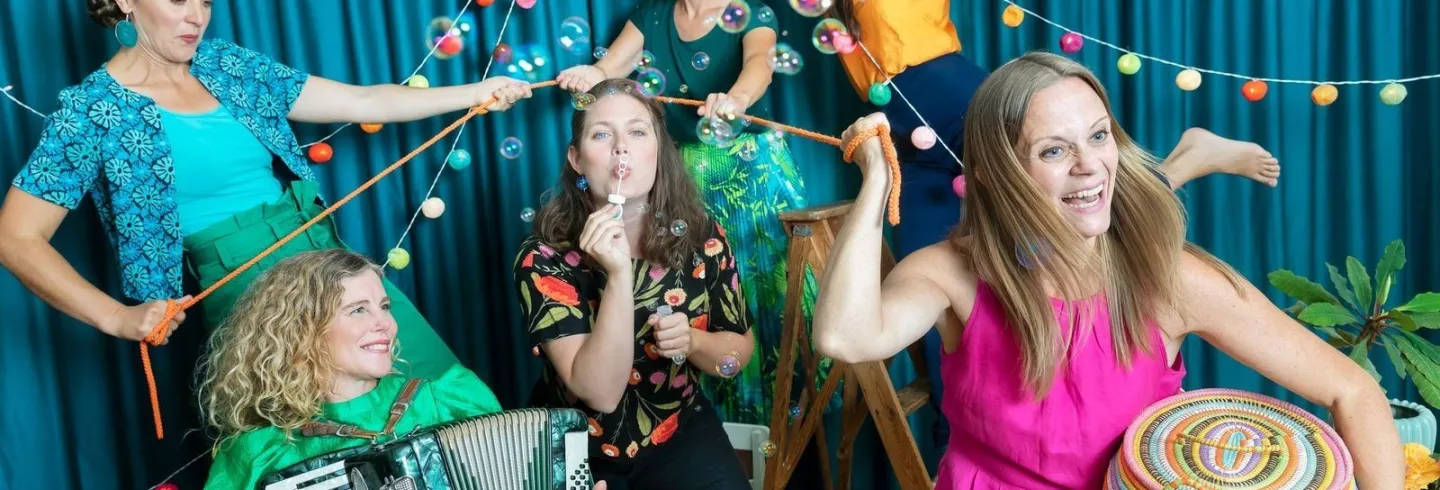 6 kvinnor i färglada kläde spelar olika instrument livligt.